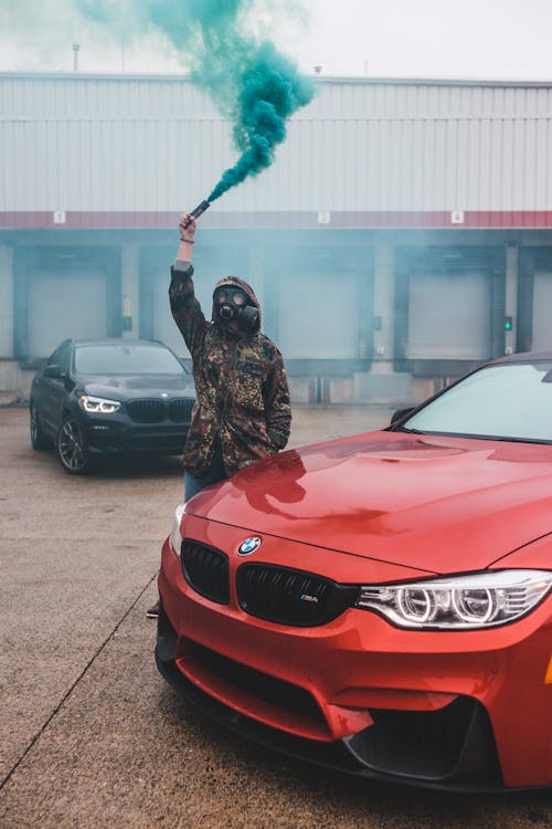 Anonymous male wearing hood and respirator standing with raised arm with green smoke flare in hand near hood of red car