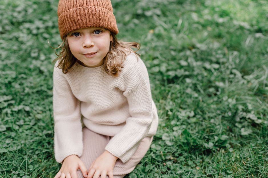 From above crop adorable girl in warm sweater and hat sitting on abundant grass in park and looking at camera contentedly