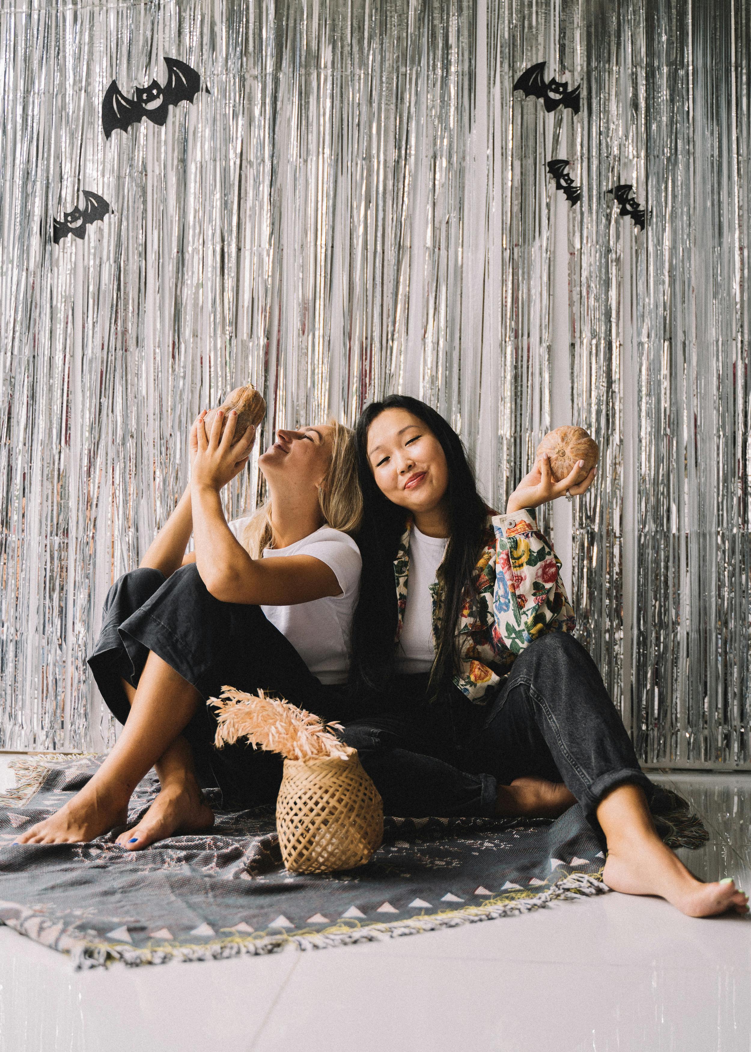 两个女人坐在室内，拿着椰子，享受着彼此的陪伴，周围装饰着万圣节的饰品