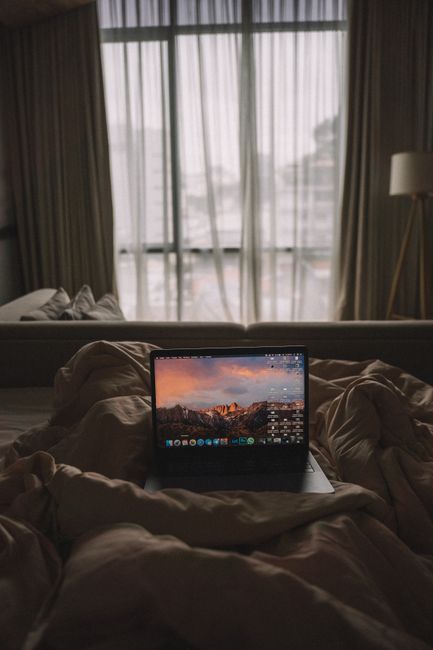 A laptop on a plush bed by the window in a cozy bedroom setting.