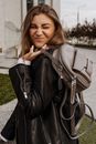 Side view young attractive female in black leather jacket making faces and carrying stylish handbag over shoulder while standing on street and looking at camera happily
