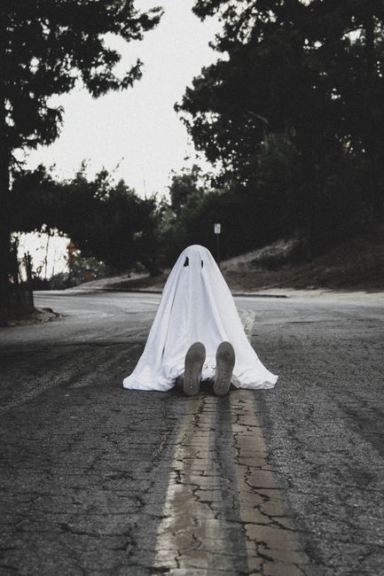 A person in a ghost costume sitting on an empty road, evoking a spooky Halloween atmosphere.