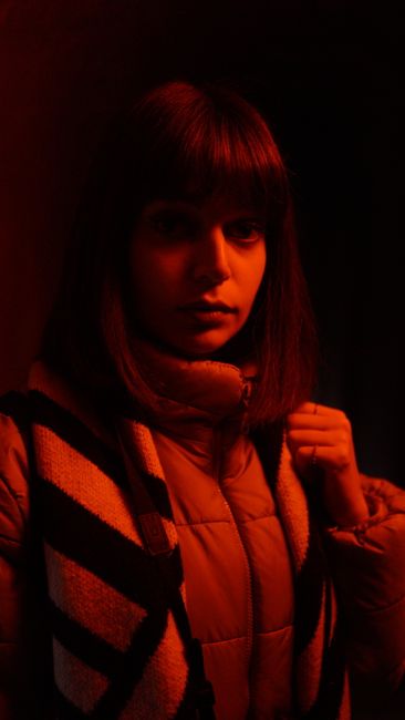 Young female wearing stylish jacket illuminated by red light looking at camera in dark room