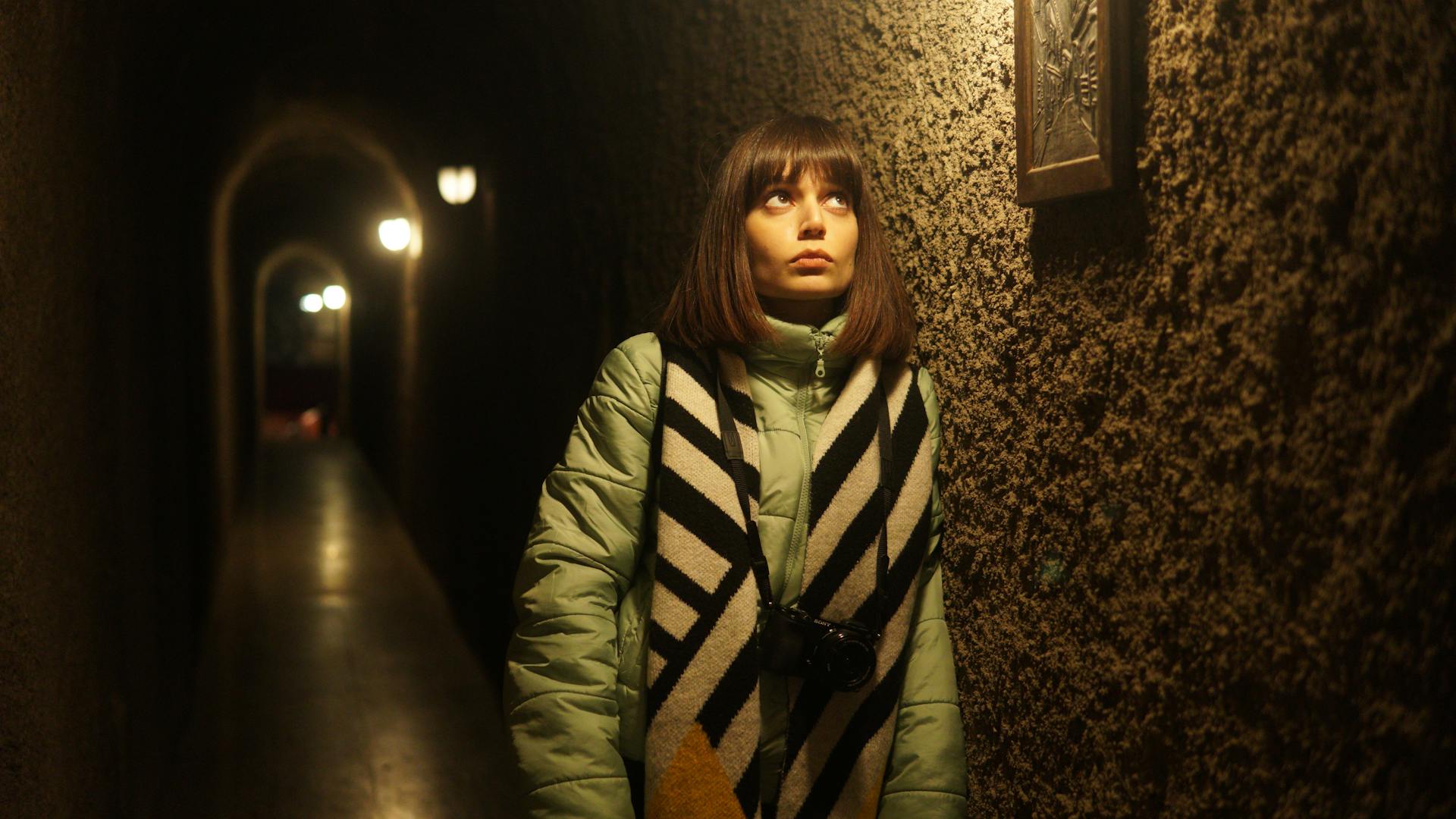 Brooding female with stylish scarf leaning on wall and looking on painting while standing in narrow corridor