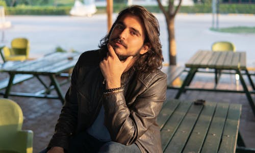 Young long haired male with beard and bracelets wearing casual clothes touching chin and looking at camera