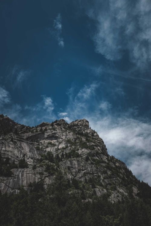 Free From below of massive rough rocky mountain with lush green trees growing on slope against cloudy blue sky Stock Photo