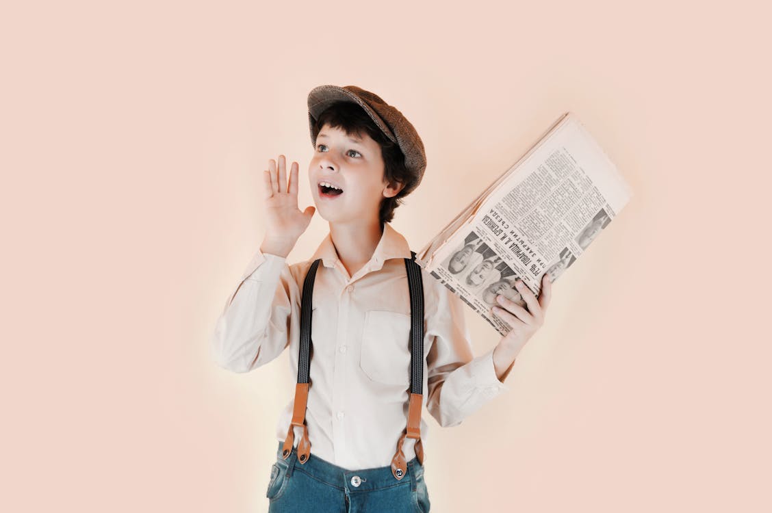 Free Positive boy in retro clothes holding old newspaper and standing with open mouth and raised arm against beige background in studio Stock Photo