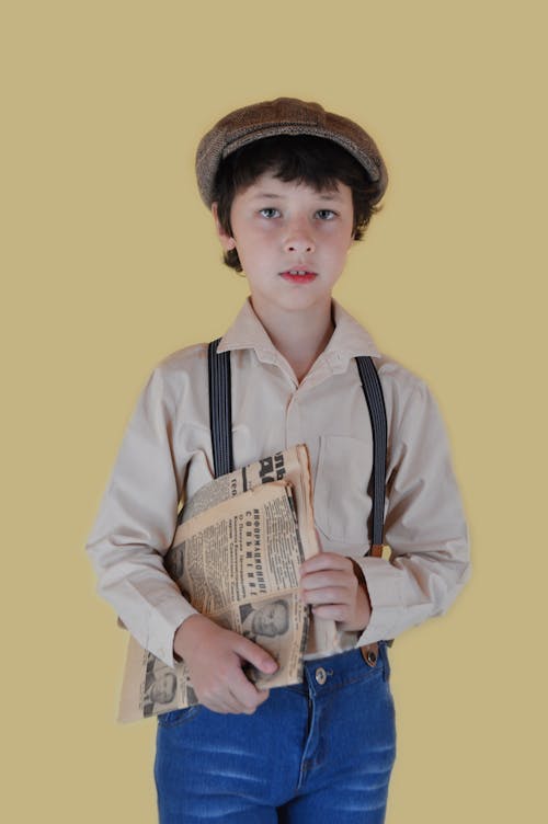 Pensive boy in old fashioned clothes standing with newspaper and looking at camera against yellow background