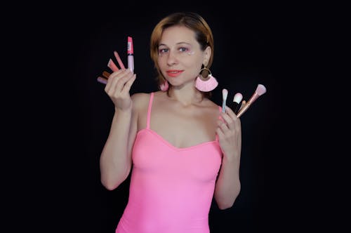 Cheerful woman makeup artist in pink top demonstrating different beauty products and cosmetic brushes and looking at camera against black background