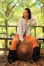 Full body of seductive woman with closed eyes and wide brimmed hat wearing fashionable blouse and colorful skirt sitting on bench in park on sunny day