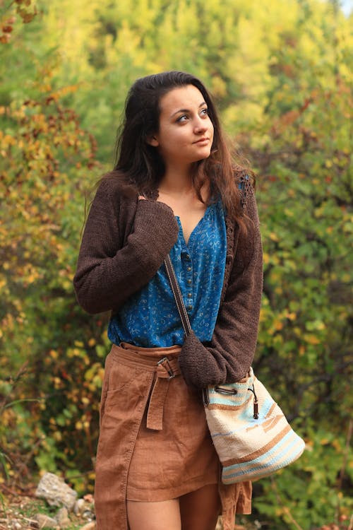 Beautiful female with handbag wearing stylish outfit looking away while standing on blurred trees background