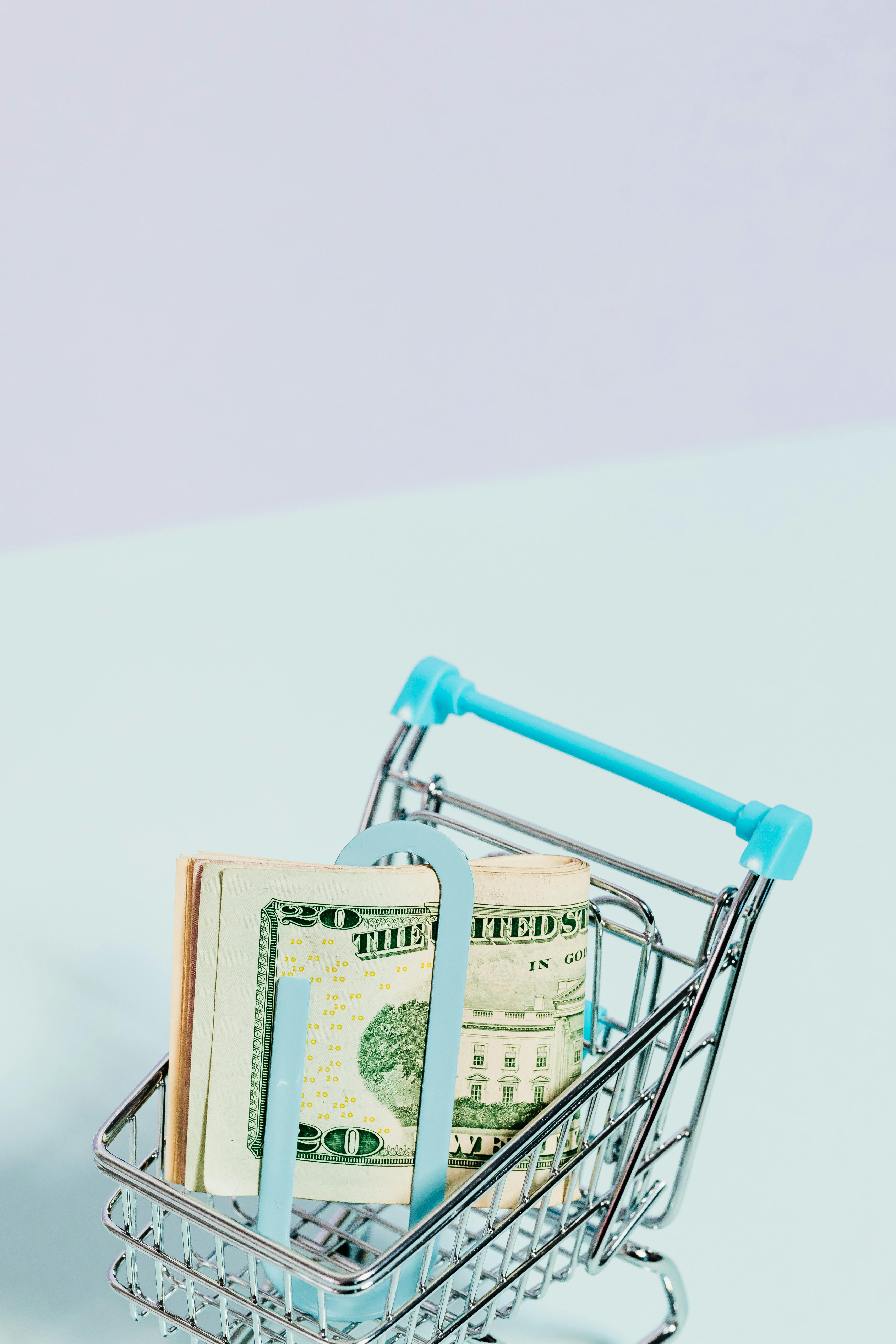 A small shopping cart filled with US dollars on a pastel blue background, symbolizing savings and spending.