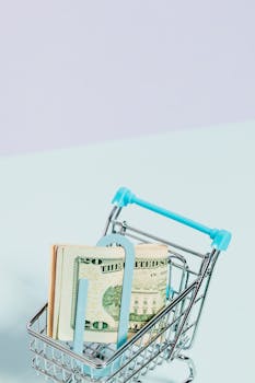 A small shopping cart filled with US dollars on a pastel blue background, symbolizing savings and spending.
