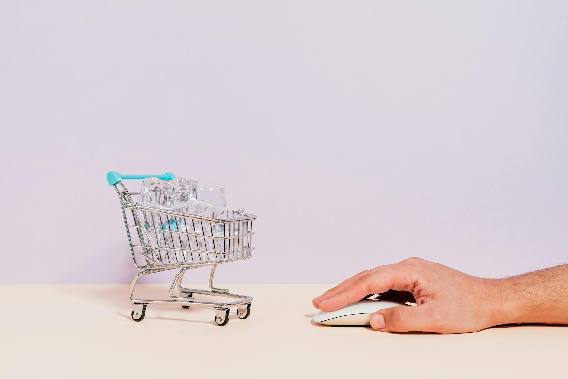 Conceptual image of a hand on a mouse next to a miniature shopping cart filled with ice cubes, symbolizing online shopping.