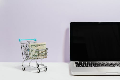 Free Macbook Pro on White Table Beside a Miniature Shopping Cart With Money Stock Photo