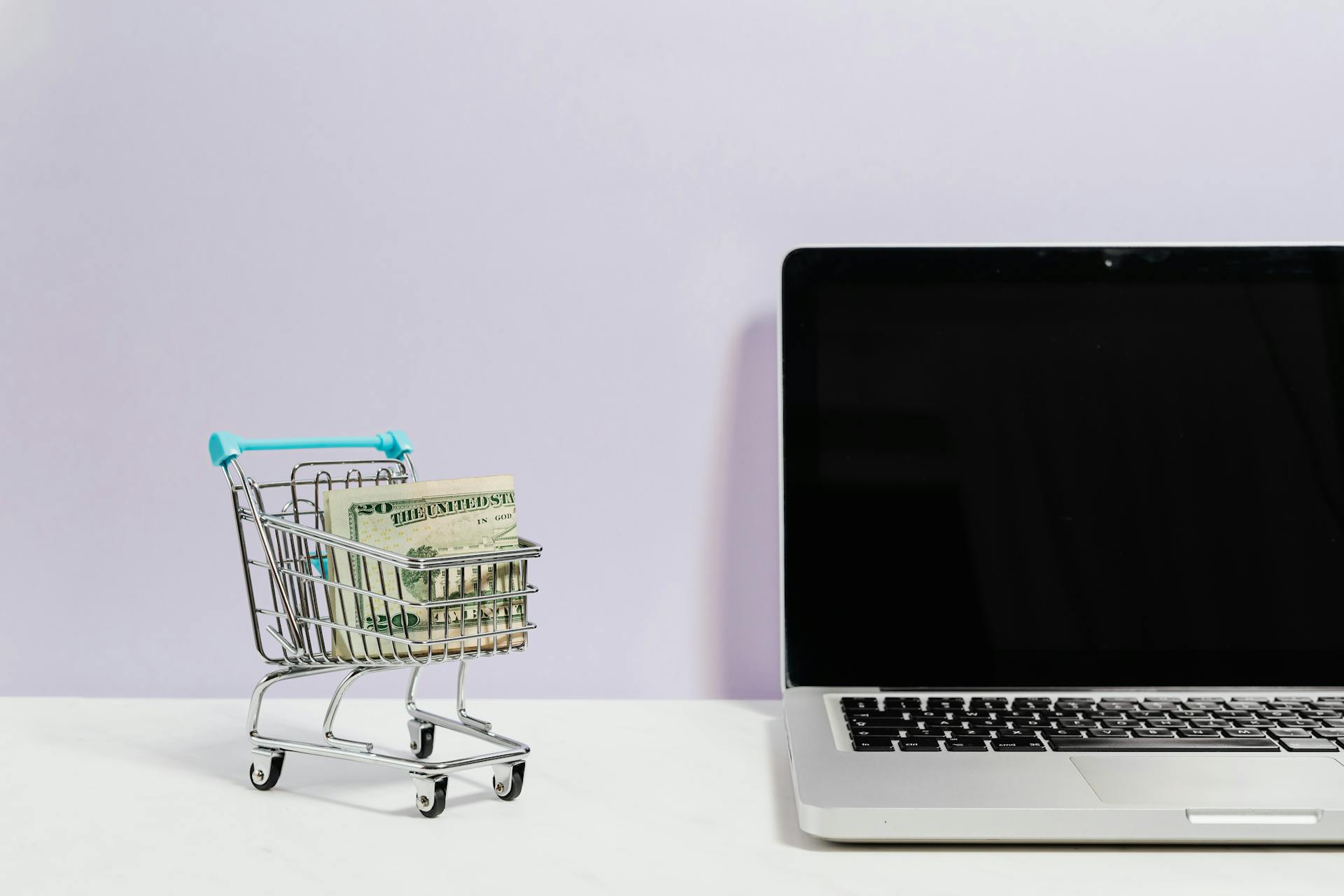 Macbook Pro on White Table Beside a Miniature Shopping Cart With Money