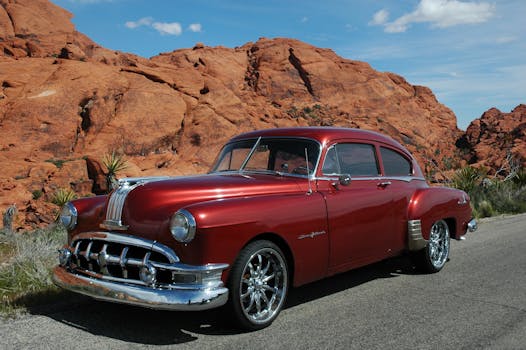 Red vintage car parked on a desert road against rocky landscape under a clear sky.