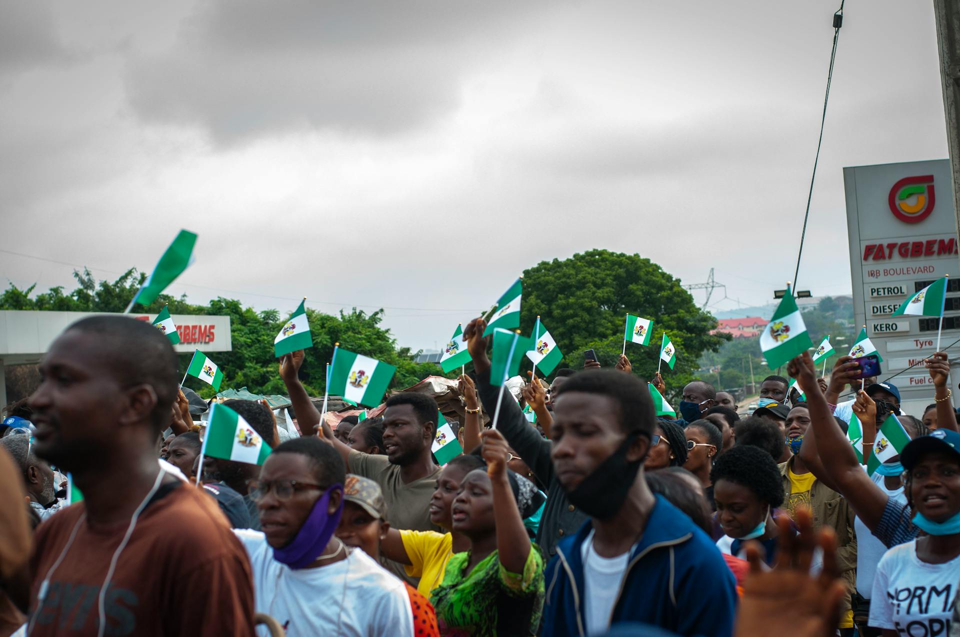 A group of people protesting in the streets, waving Nigerian flags under a cloudy sky.