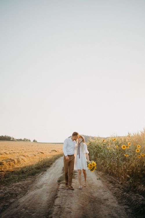 Free A couple walking on a dirt path surrounded by sunflowers under a clear sky, depicting love and tranquility. Stock Photo