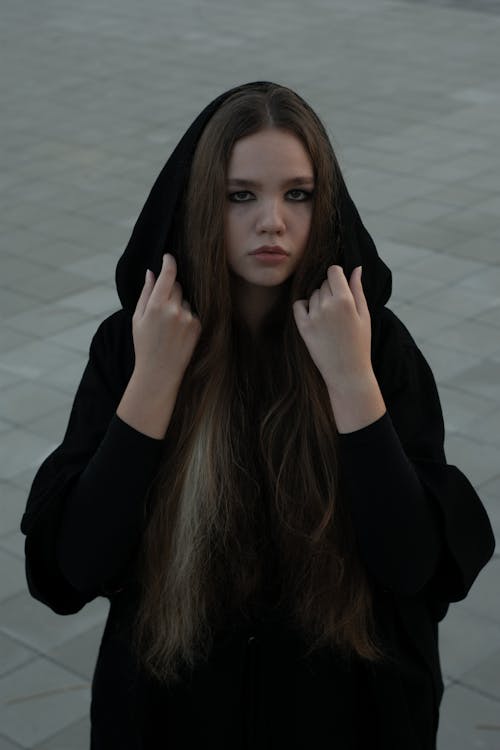 From above sad solemn female wearing black loose clothes and touching hood while standing on paved street and looking at camera