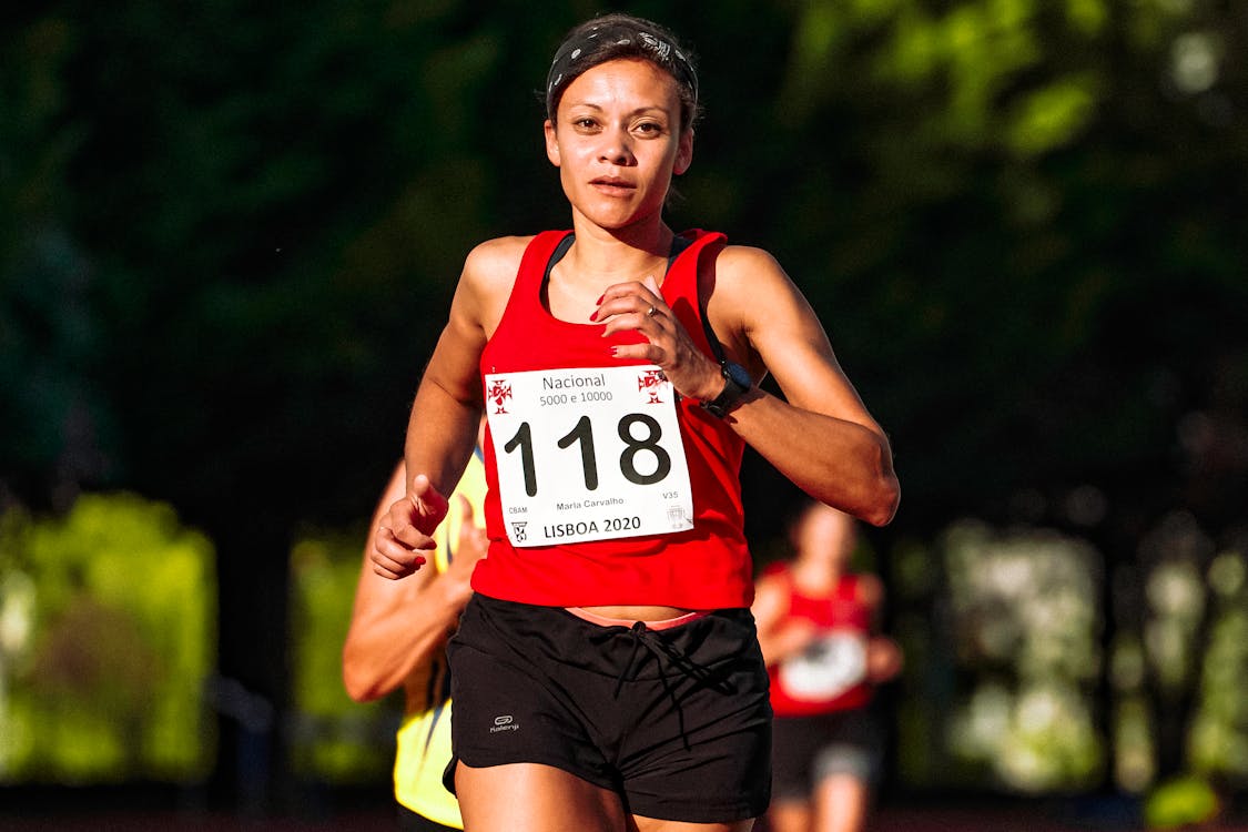 Determined ethnic sportswoman in active wear with numbers running during track and field competition while looking at camera