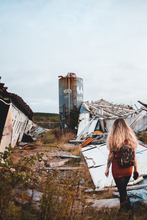 Free Back view of anonymous female with backpack strolling along damaged wall with remains of broken construction with rusty tank in distance Stock Photo