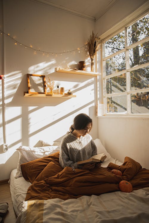 Woman in Gray Long Sleeve Shirt Sitting on Her Bed Reading a Book