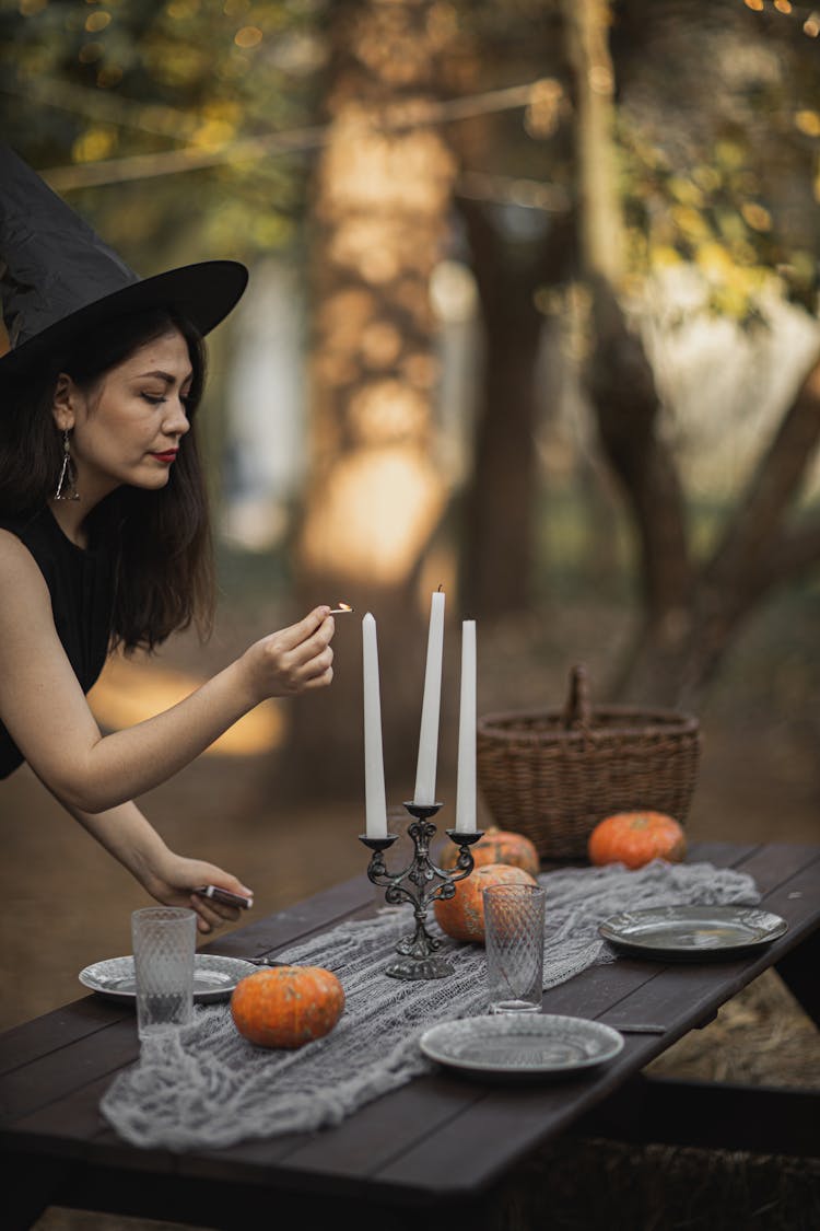 Woman In Black Witch Costume Lighting A Candle On Table