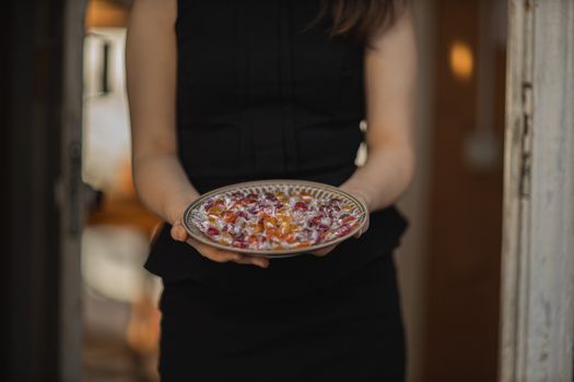 一名身穿黑色连衣裙的女子端着一盘装满彩色万圣节糖果的盘子，非常适合用来玩“不给糖就捣蛋”