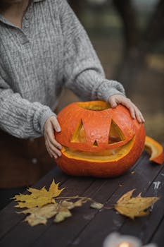 Person carving a smiling pumpkin outdoors surrounded by autumn leaves, perfect for fall celebrations.
