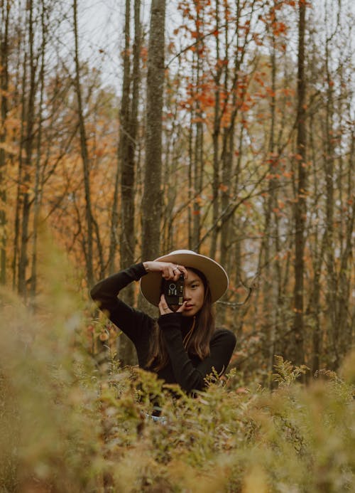 Concentrated young ethnic female traveler in elegant outfit and hat taking photo on retro film camera while relaxing in picturesque autumn forest