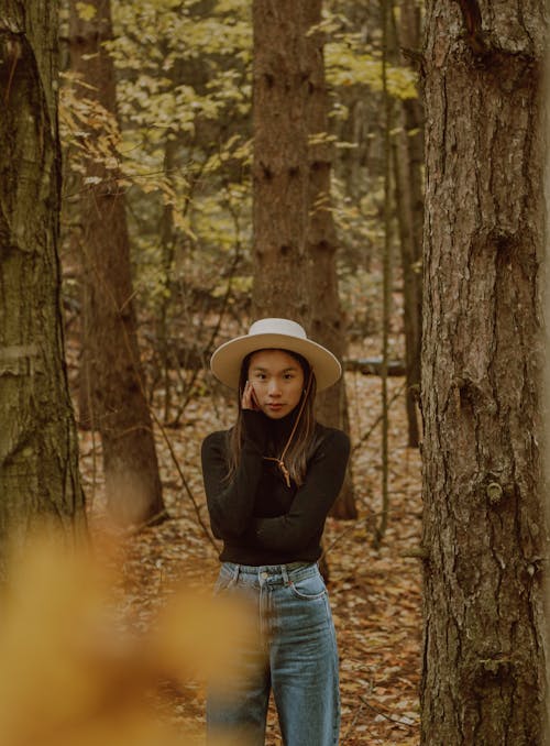 Fashionable young ethnic woman in stylish outfit and hat touching cheek and looking at camera in autumnal park