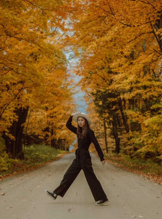 Woman in Black Jacket and Black Pants Walking on Road Between Trees