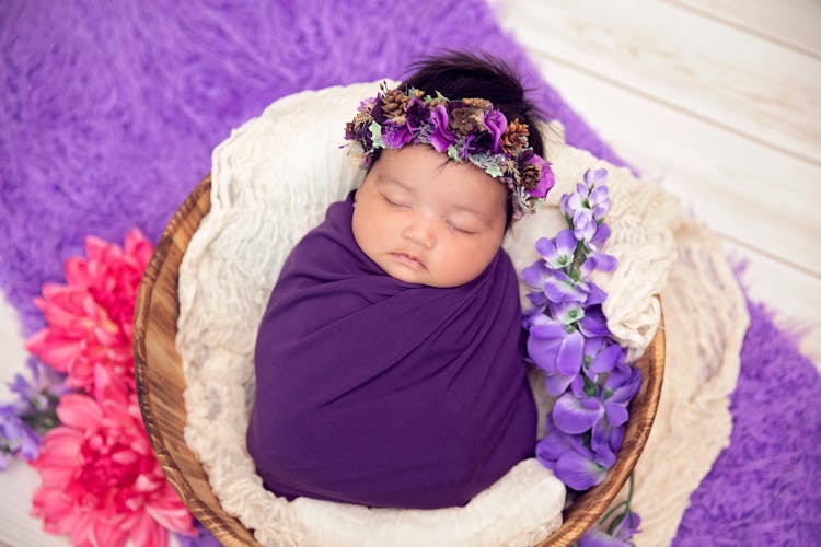 A Cute Little Baby In Purple Swaddle Lying On A Basket