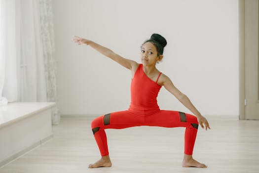 A young girl in a red outfit performs a gymnastics pose in a well-lit indoor space.