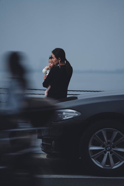 Back view calm young female in black wear holding little baby on hands while standing embankment near moving cars