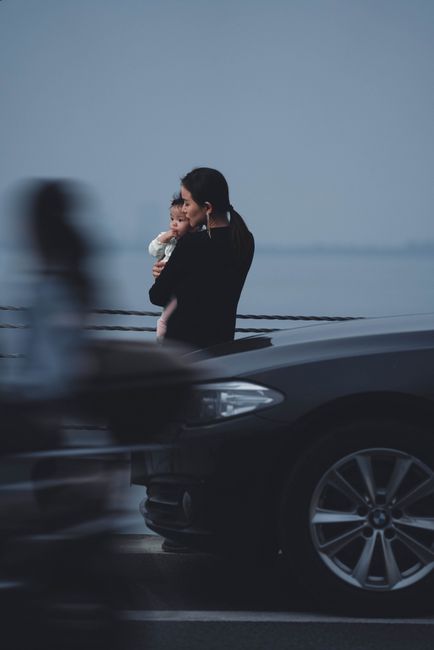 Back view calm young female in black wear holding little baby on hands while standing embankment near moving cars