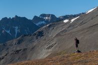 Man in Black Jacket Standing on Green Grass Field Near Gray and White Mountain