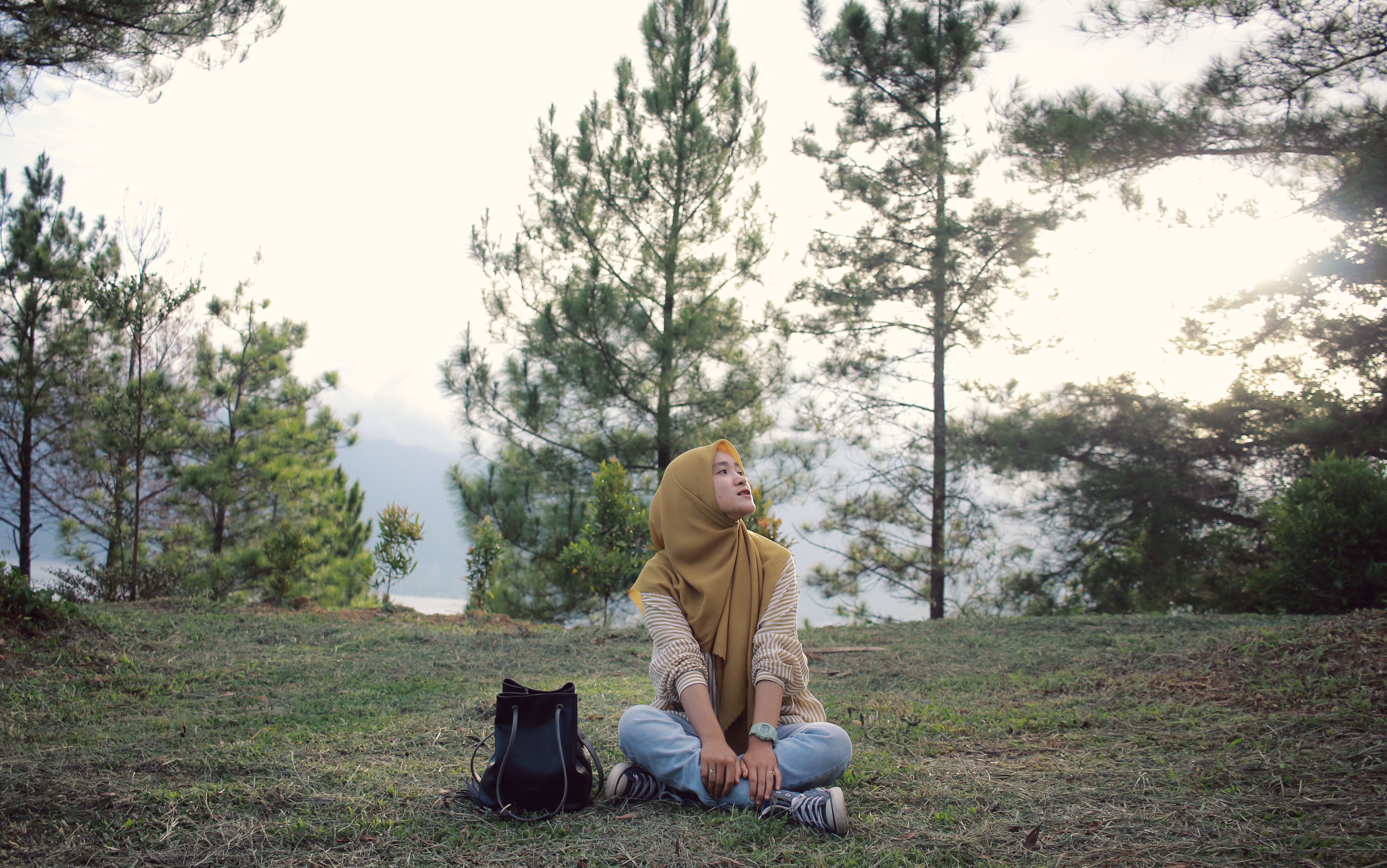 Young woman with hijab sits on grass in a forest, looking contemplative.