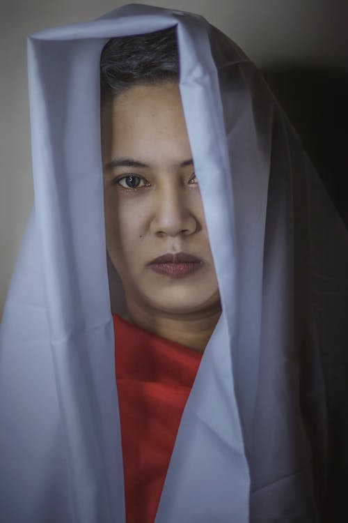 Serious ethnic young lady covering head with white cloth and covering breast with red fabric while looking at camera in light studio