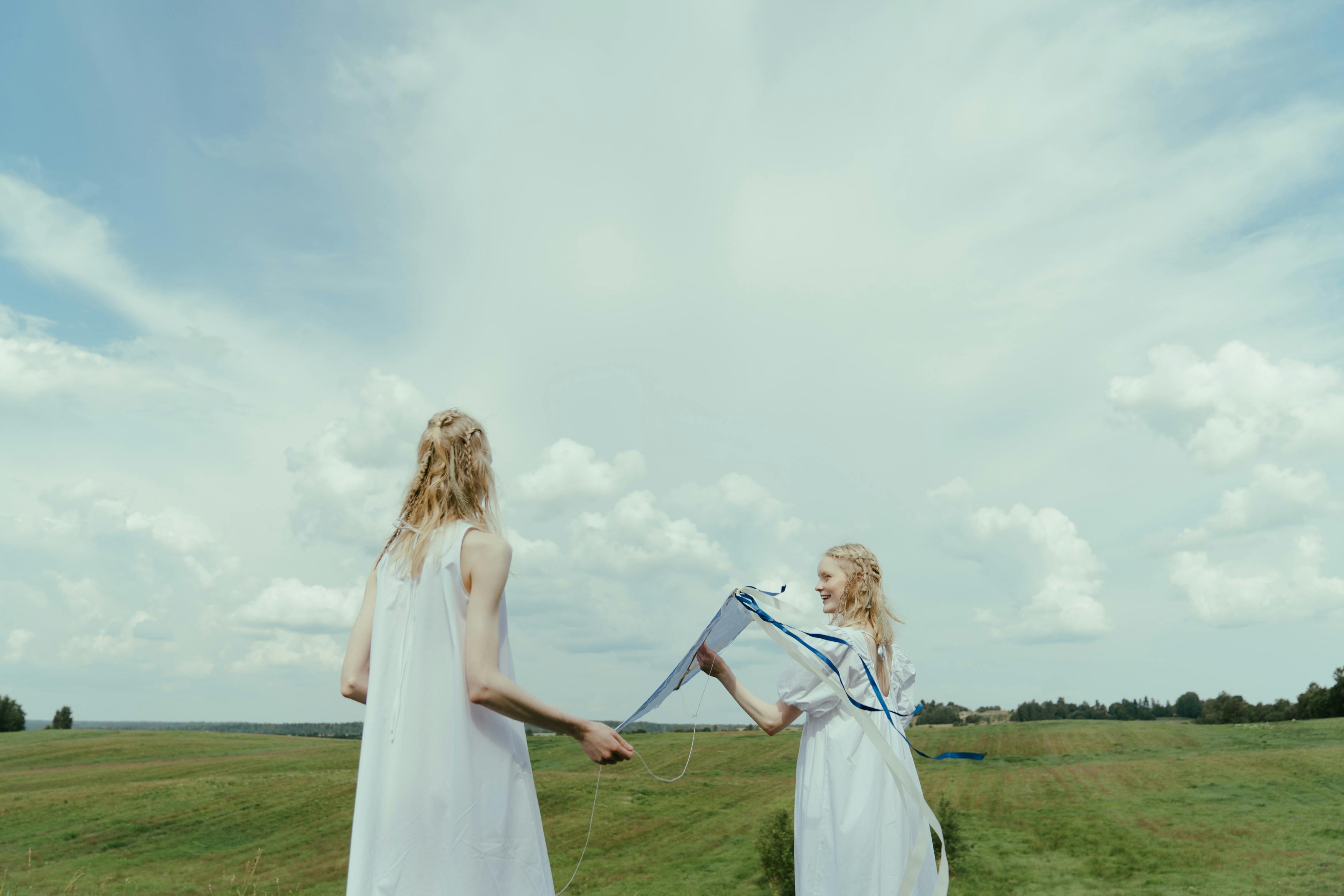Two women enjoy a summer day flying a kite in a wide, grassy field.