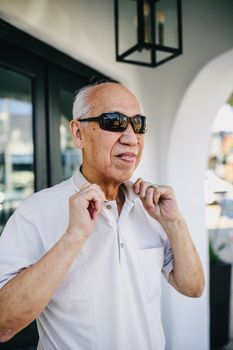 阳光明媚的一天，一位戴着太阳镜的亚洲高级男子在大楼外整理衣领