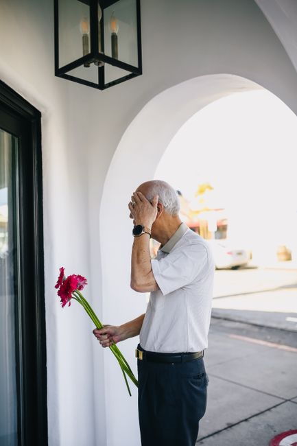 Senior man holding red flowers, standing by a modern archway outdoors, appearing thoughtful.