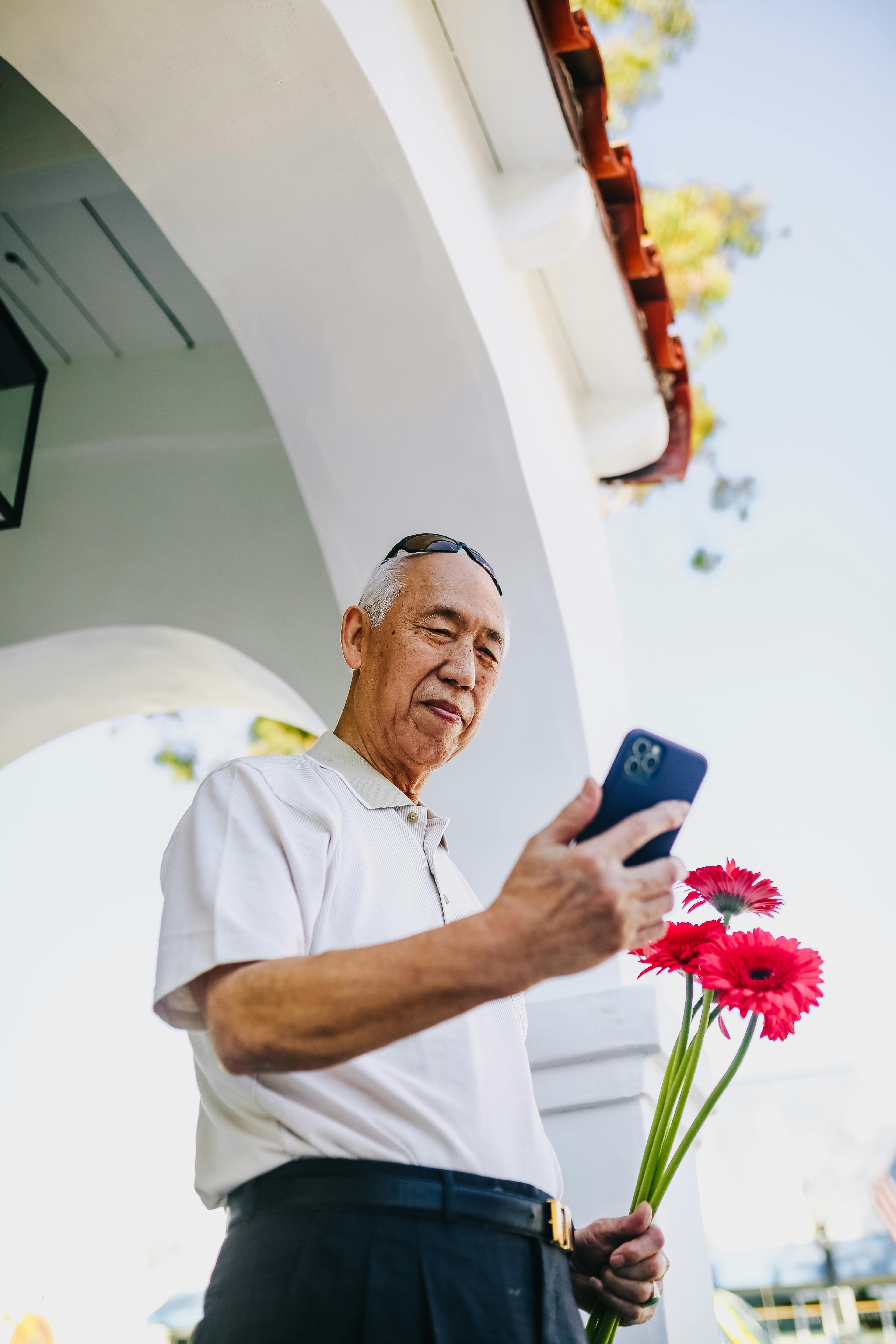 Elderly man holding flowers and smartphone, standing under bright arch outdoors.