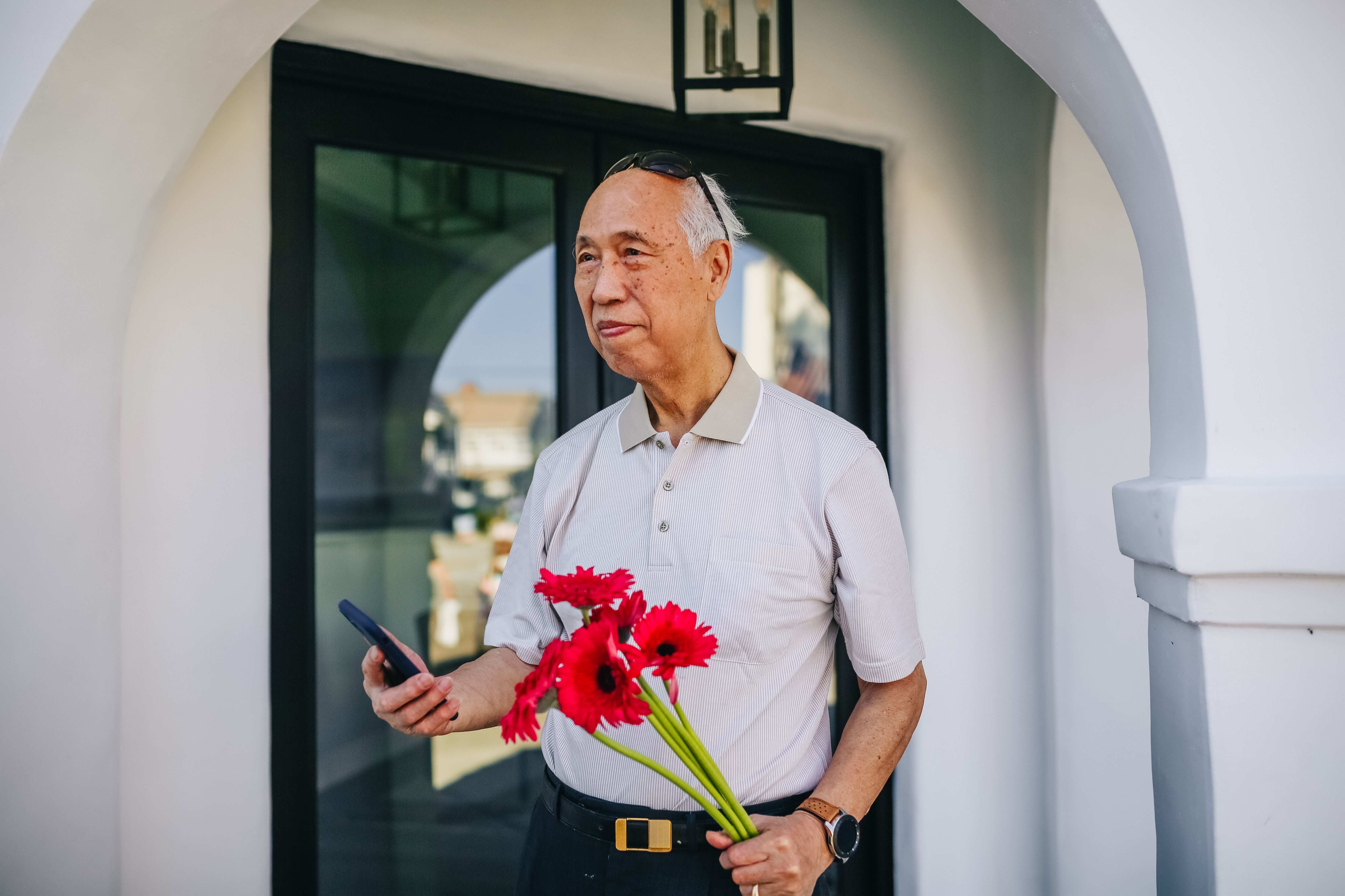 一位亚洲老人手持一束红花和一部智能手机，站在建筑物外