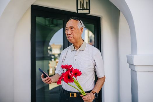 一位亚洲老人手持一束红花和一部智能手机，站在建筑物外
