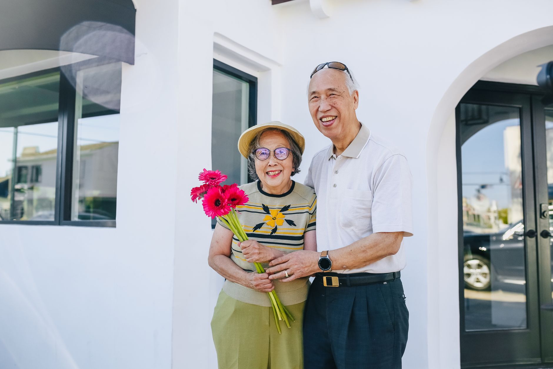 Smiling senior couple holding flowers on a sunny day outside a building.