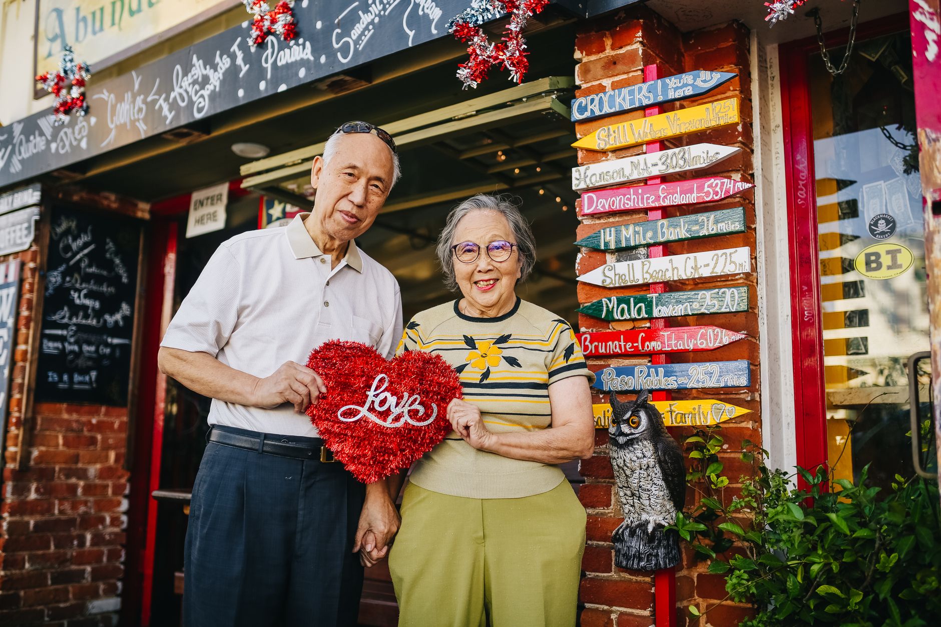 Senior couple holding a heart symbol outside a colorful shop, showcasing love and joy.