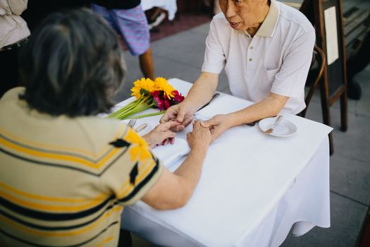 一对亚洲老年夫妇在户外咖啡馆手牵着手，分享着温暖的亲密时刻，画面十分迷人