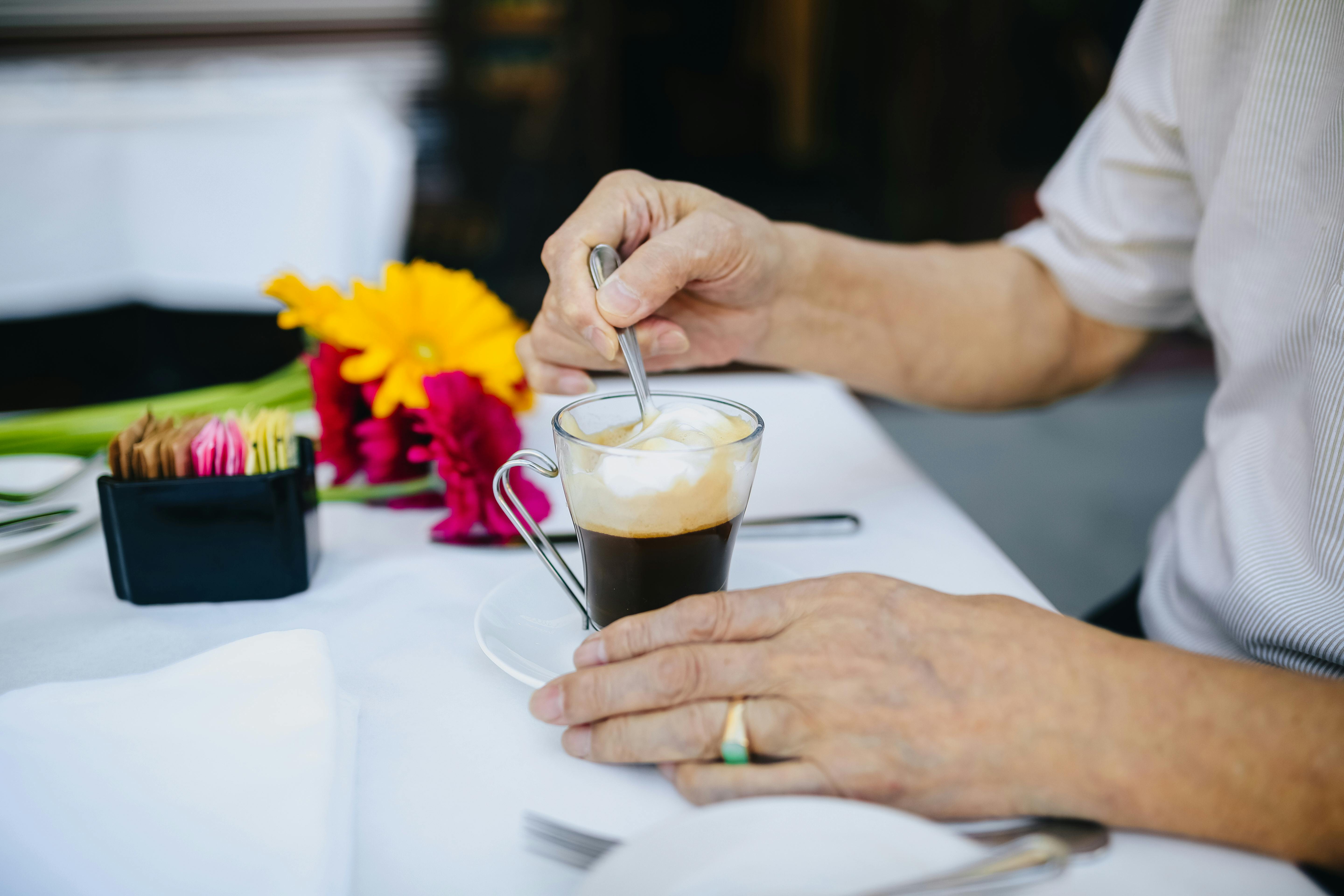 一位年长男子在户外咖啡馆的桌边搅拌着卡布奇诺，身旁盛开着鲜艳的鲜花